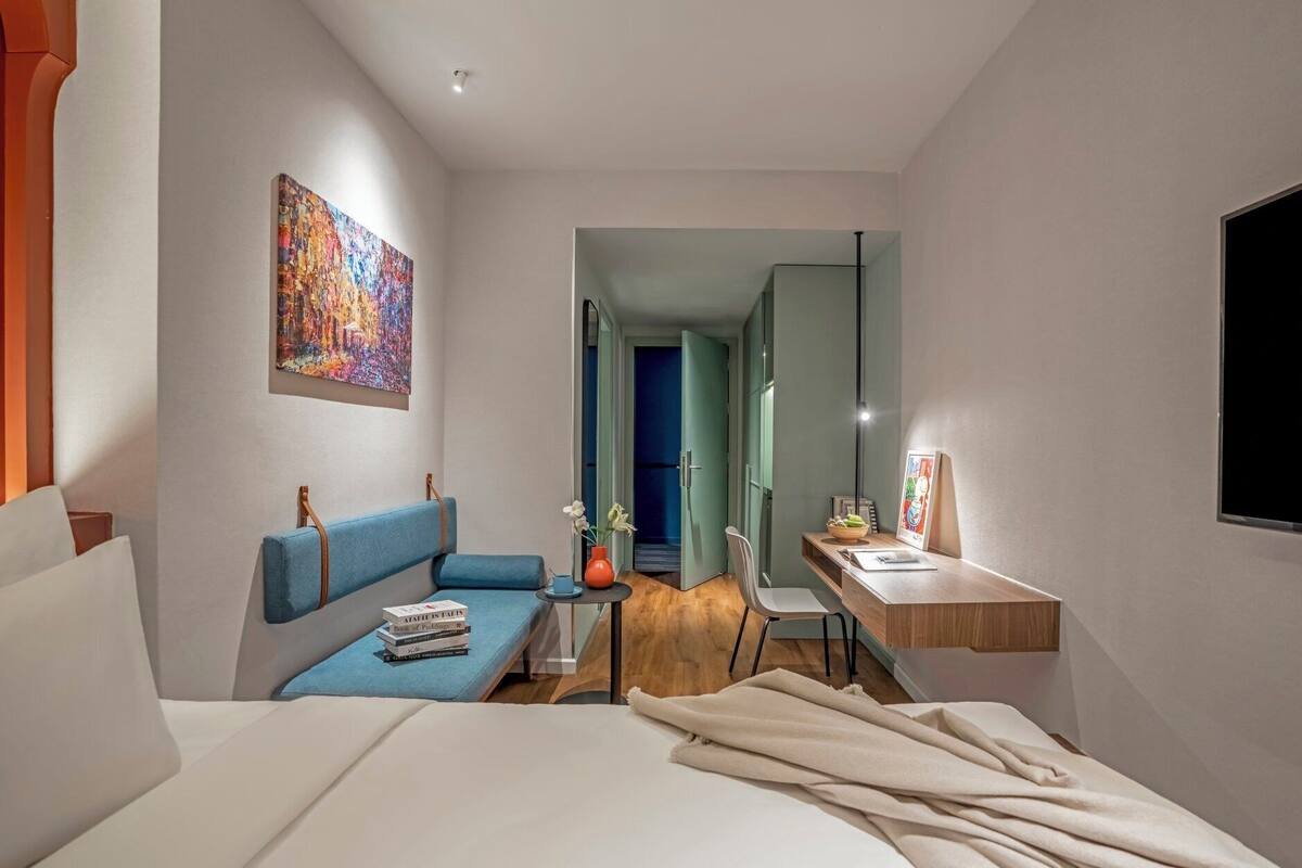 Une chambre d’hôtel moderne avec un lit, un canapé bleu, une petite table avec un vase et un bureau avec une chaise.