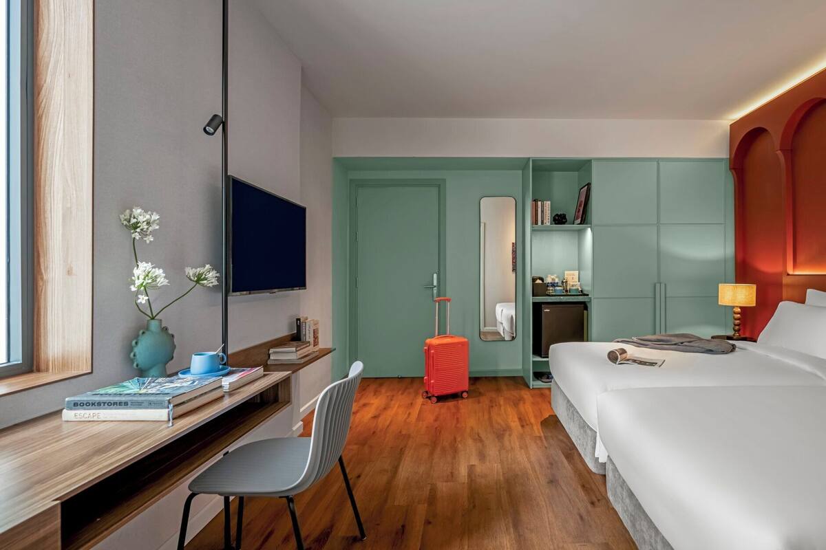 Une chambre d’hôtel comprenant un lit, un bureau avec une chaise, une télévision, un miroir, une armoire et une valise.