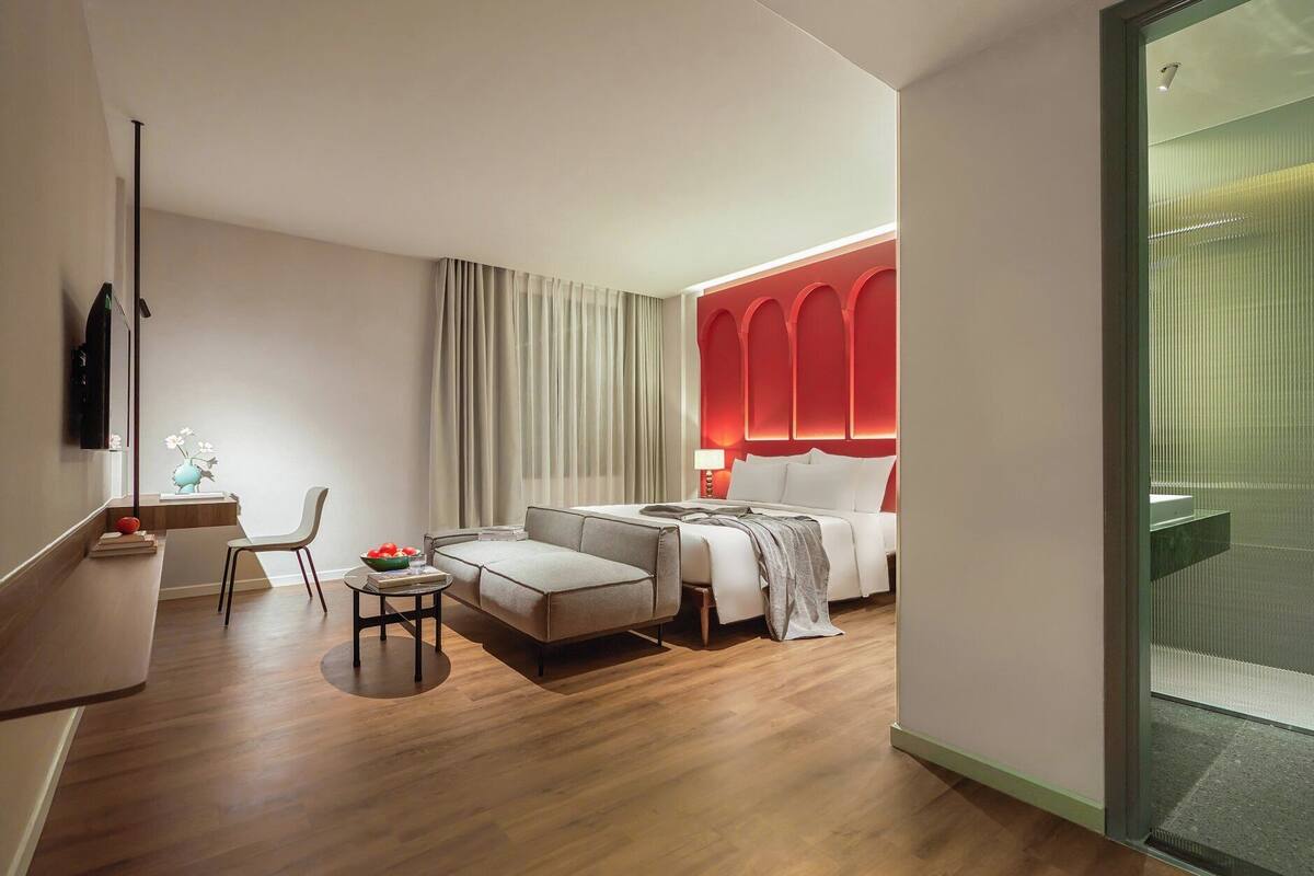 Une chambre d’hôtel moderne comprenant un lit, un coin salon avec une table et des chaises, un petit bureau avec une lampe et une grande fenêtre avec des rideaux.