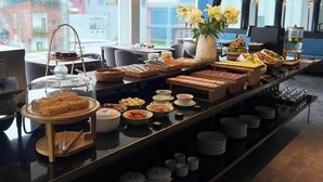 A buffet setup with various food items and a coffee station, set in a modern dining area with large windows and decorative lighting. - M Village Hotel Đà Nẵng Centre, a brand of Modern Village Lifestyle (Da Nang)