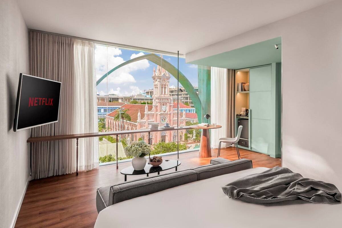 Une chambre d’hôtel moderne dotée d’une grande fenêtre donnant sur un bâtiment historique. Un téléviseur à écran plat fixé au mur affiche le logo de Netflix.