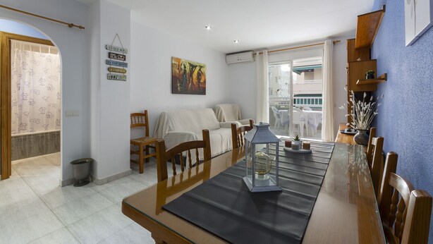 A dining area with a long wooden table, chairs, and a glass-top table. There is a small trash bin and a window with curtains. - AZUL - Cosy flat with shared swimming pool and free wifi (Daimús)