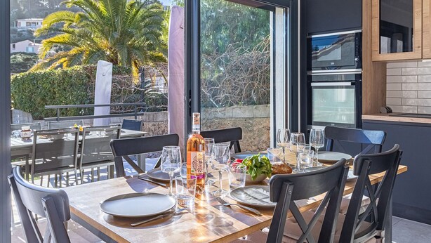 A dining area with a table set for four, large windows offering a view of palm trees and a hillside, and a modern kitchen with dark cabinetry and stainless steel appliances. - Maison Fabrégas by Interhome (La Seyne Sur Mer, Les Sablettes)