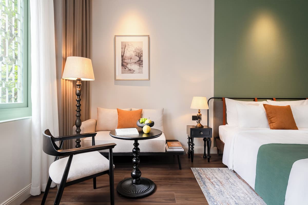 Habitación de hotel con cama, sofá, una mesita con lámpara, una silla y un cuadro en la pared.