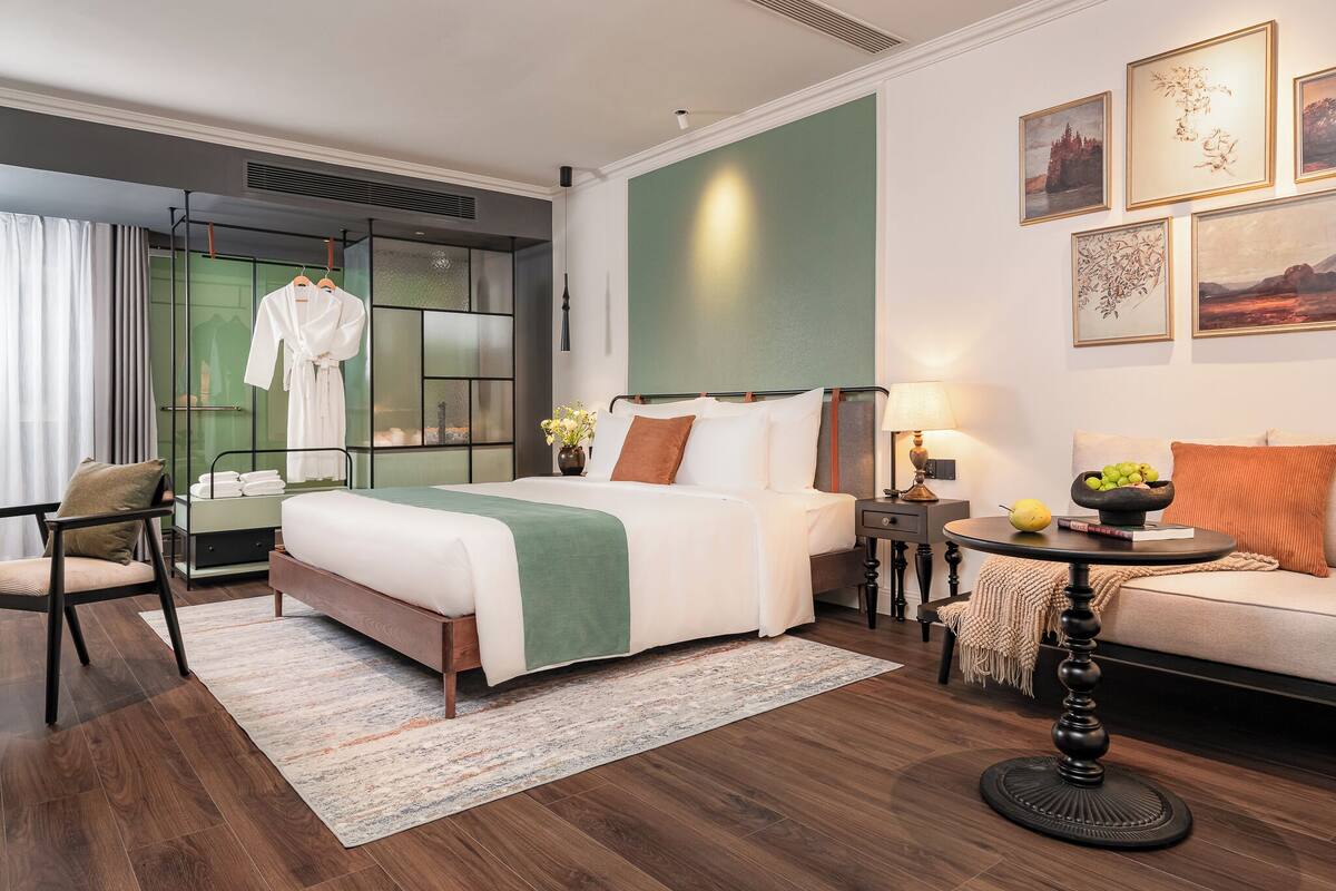 Habitación de hotel moderna con una cama grande, un sofá, una mesita y una silla. Se ve un albornoz en un perchero, un jarrón con flores y una obra de arte en la pared.