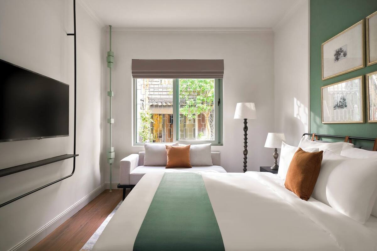 Habitación de hotel con cama, televisor, ventana con vistas a follaje y cuadros en la pared.