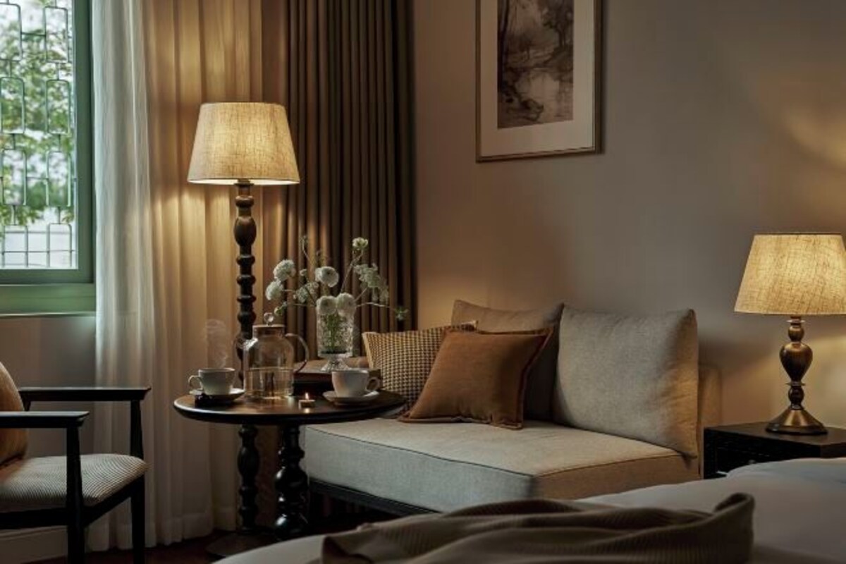 A hotel room with a sofa, two lamps, a small table with a cup and saucer, and a vase with flowers.