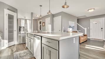 A modern kitchen with stainless steel appliances, a large island, pendant lighting, and a view to the living area.