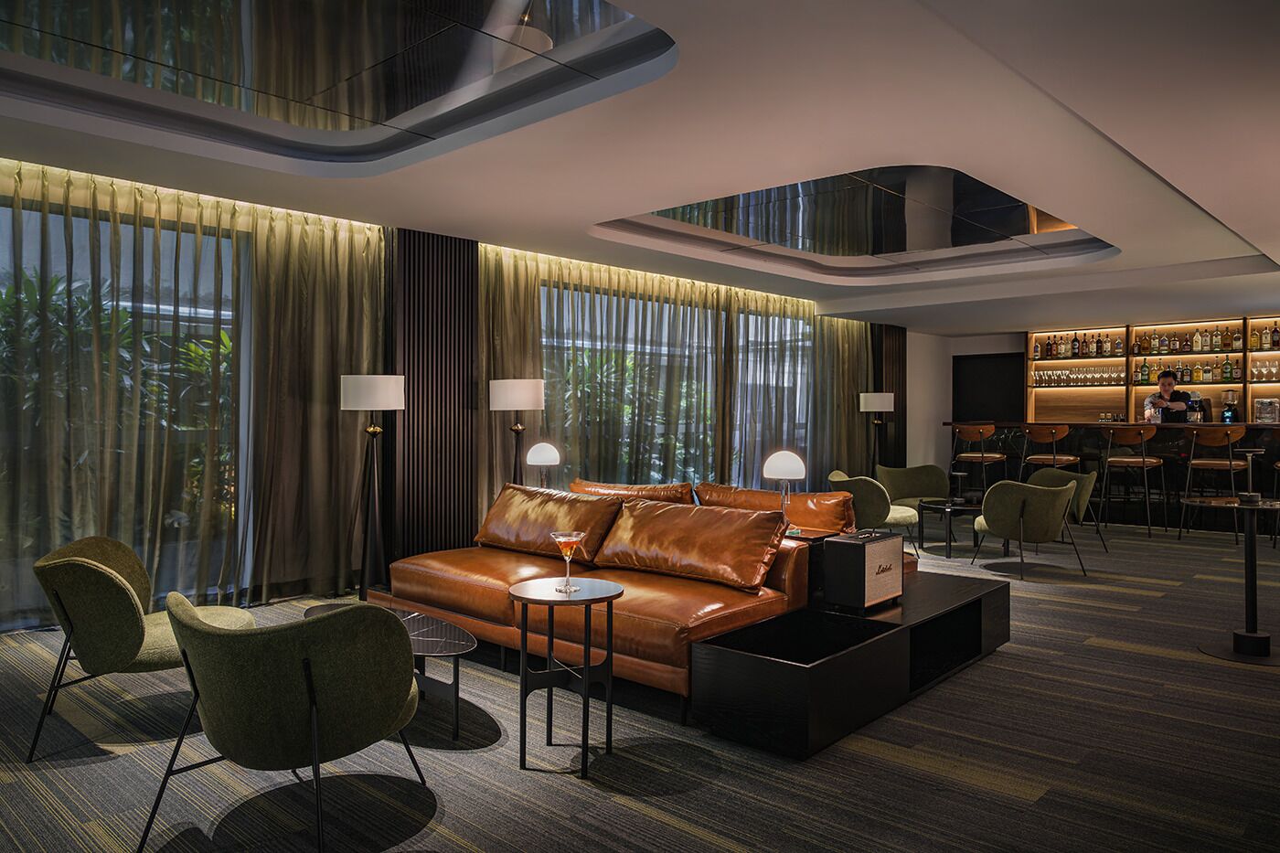Un hall d’hôtel moderne avec un canapé en cuir, des fauteuils verts, un coin bar et de grandes fenêtres avec des rideaux.