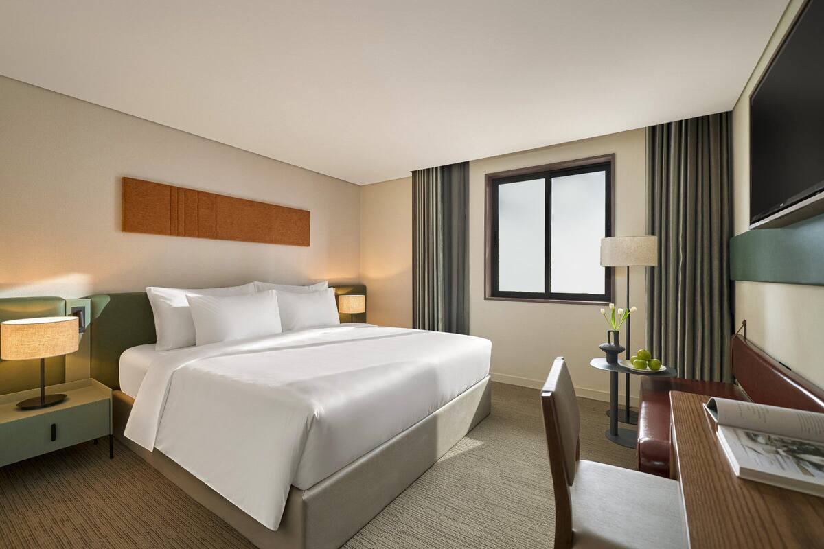Une chambre d’hôtel comprenant un lit, des tables de chevet, un bureau avec une chaise, une télévision et une fenêtre avec des rideaux.