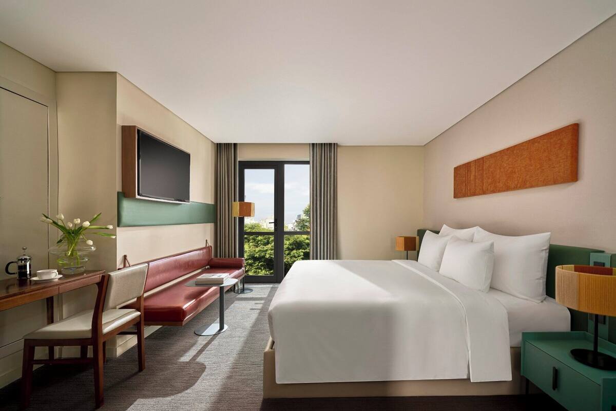 Une chambre d’hôtel comprenant un lit, un bureau avec une chaise, un banc, une télévision et une fenêtre donnant sur un paysage verdoyant et des bâtiments.