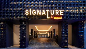 Das moderne Hotel zeigt sich mit einer geometrischen Fassade und leuchtenden Hinweisschildern mit dem Schriftzug „SIGNATURE BY M VILLAGE“.
