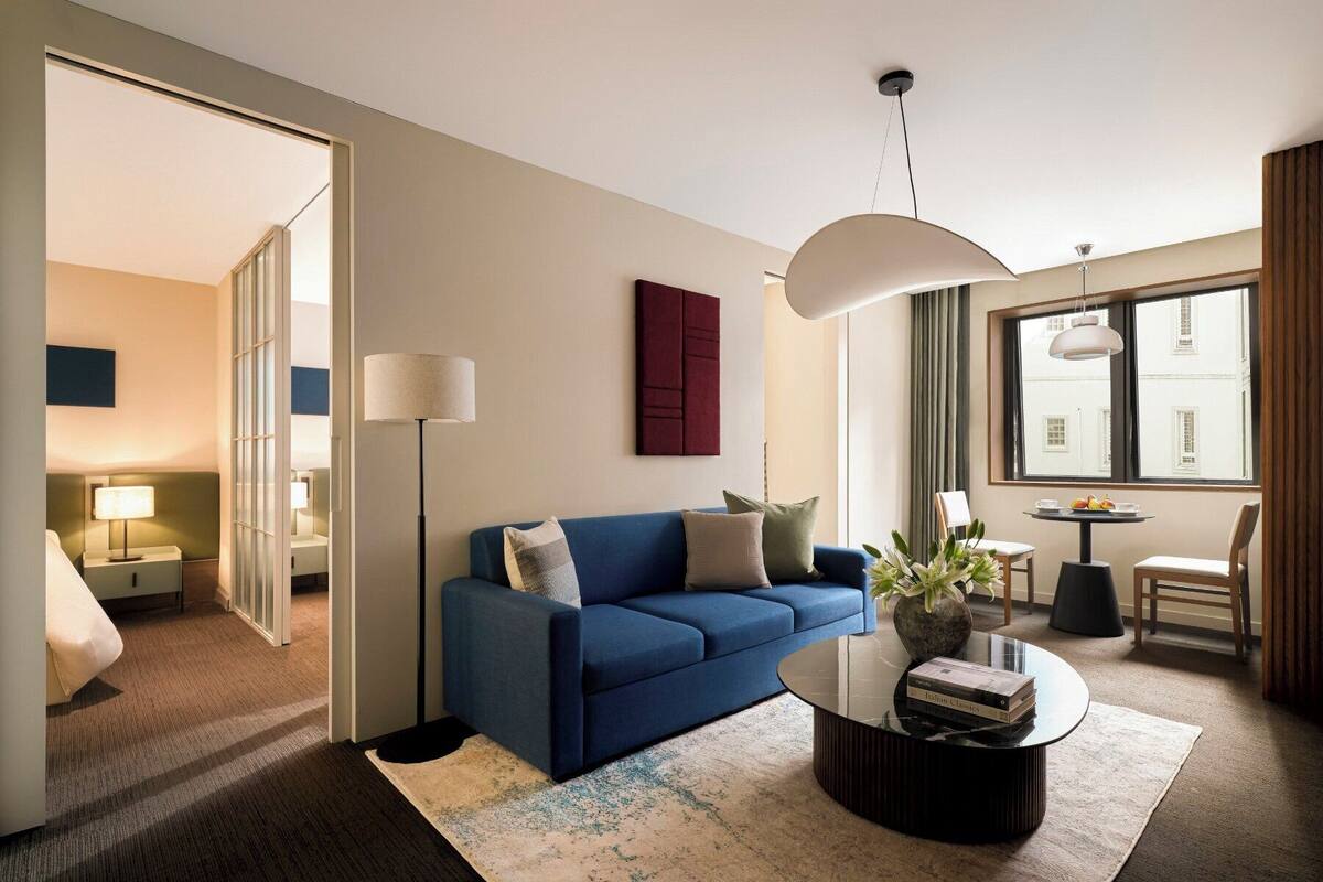 Un salon moderne avec un canapé bleu, une table basse ronde et une grande fenêtre donnant sur des immeubles.
