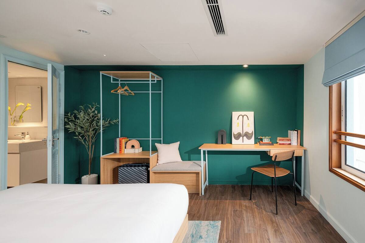 A modern hotel room with a bed, a desk, a chair, a hanging clothes rack, a small plant, and a window with a view.