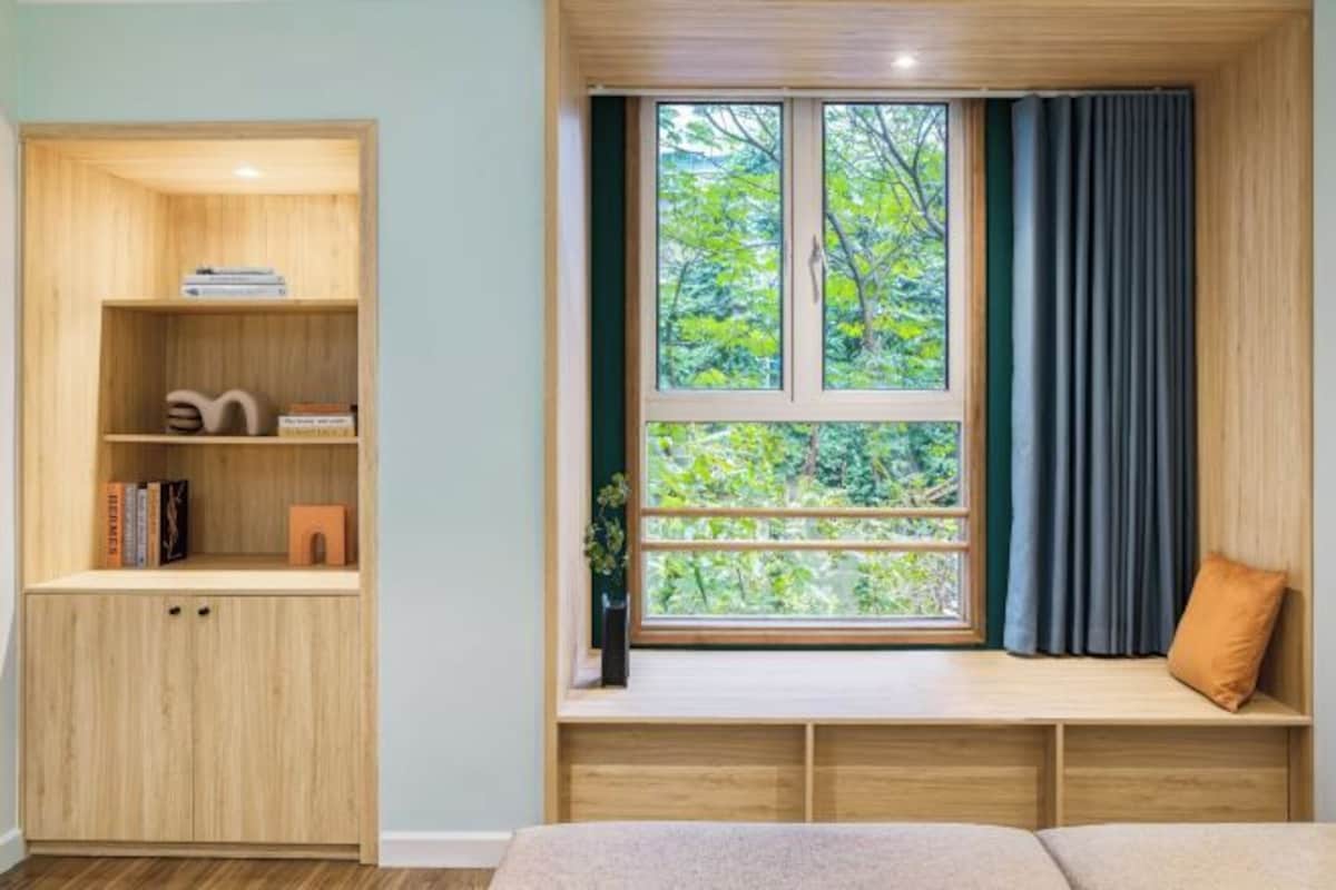 A room with a built-in wooden bookshelf, a window with a view of greenery, and a built-in bench with a cushion.