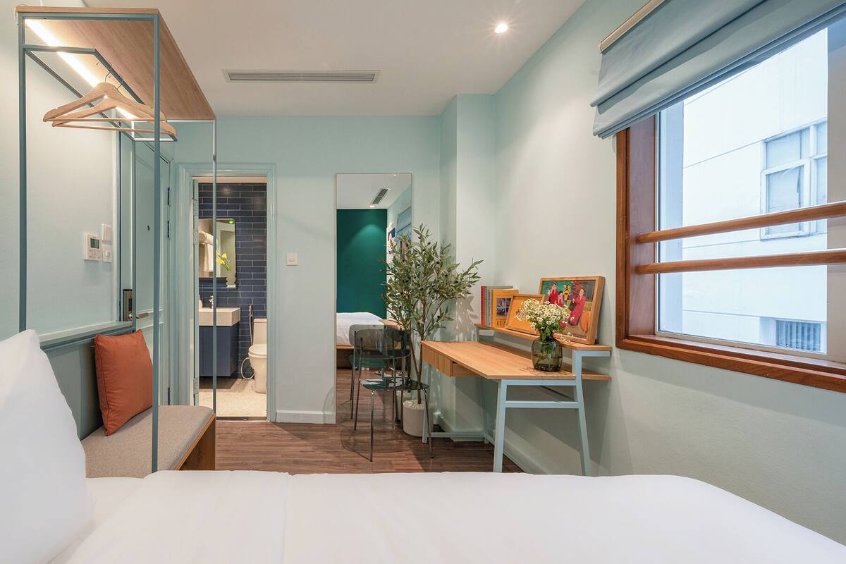 A hotel room with a bed, a desk, a chair, a window, and a bathroom visible through an open door.