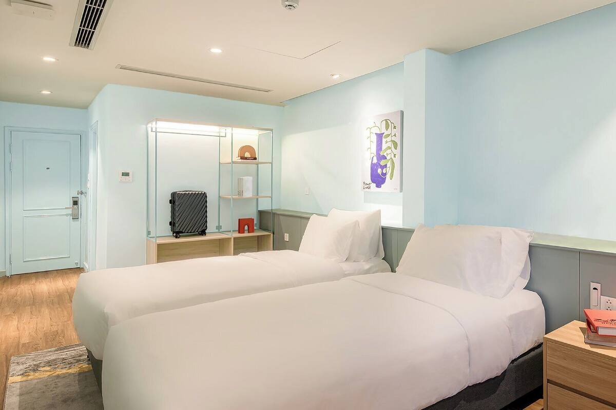 A hotel room with two beds, a wardrobe, a mirror, a shelf with books, and a wall art piece.