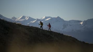 Deux personnes font de la randonnée sur une pente herbeuse, avec des montagnes enneigées en arrière-plan.