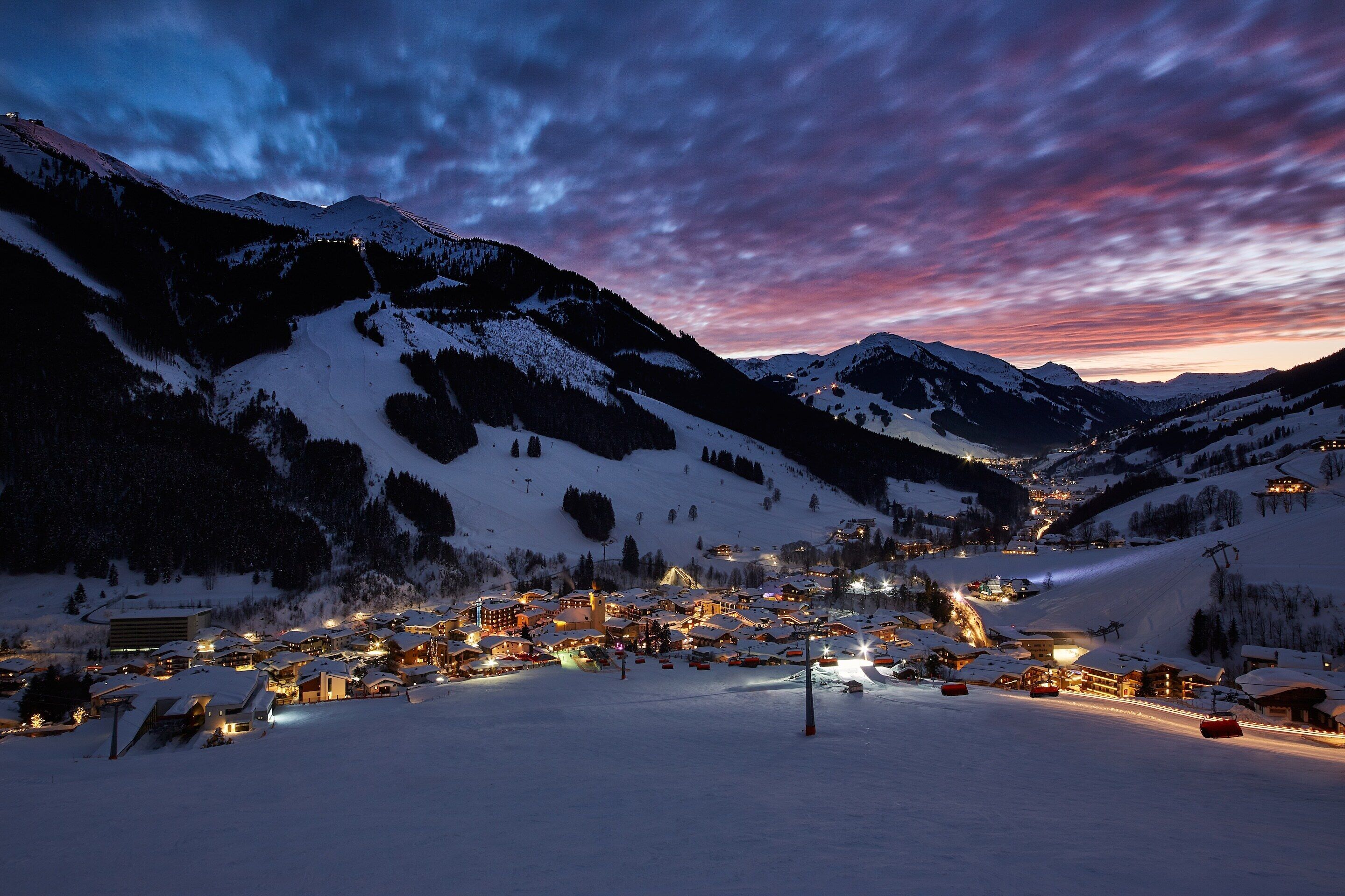 A snow-covered mountain town at dusk with illuminated buildings and a ski slope.