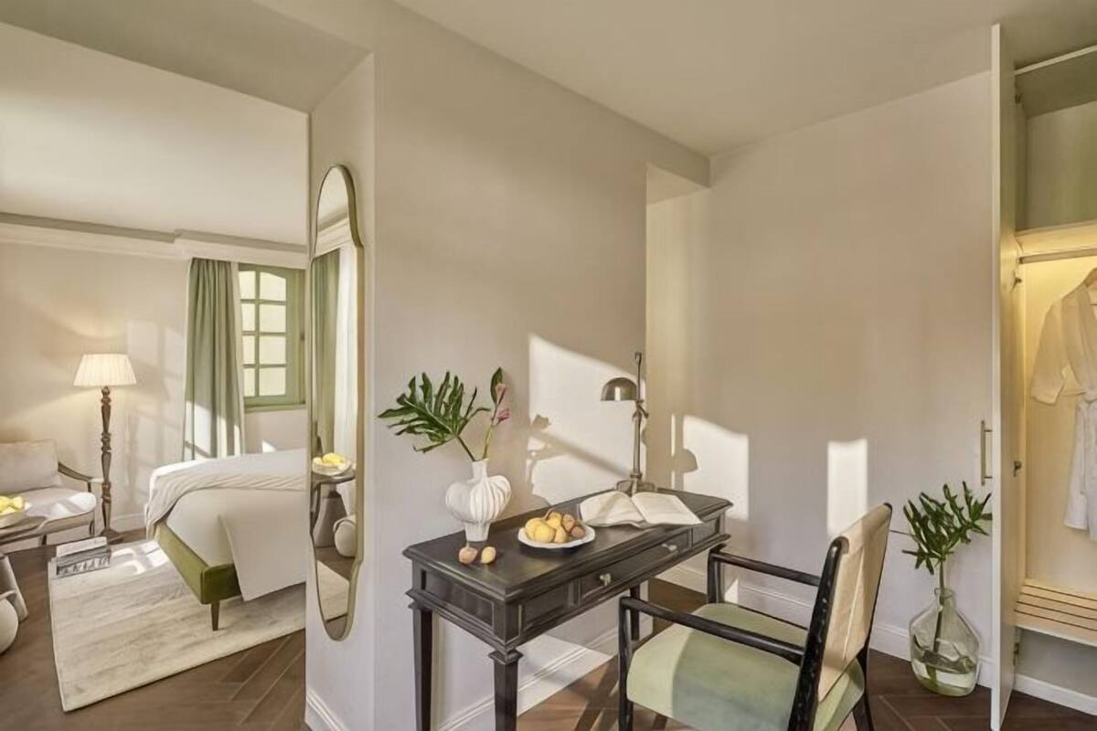 A hotel room with a bed, bedside table, lamp, mirror, and a desk with a chair.