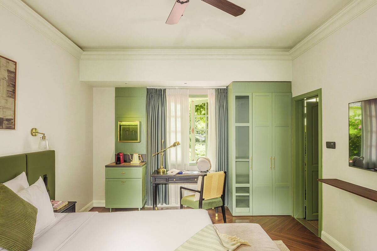 A hotel room with a bed, desk, chair, and a view of greenery outside.