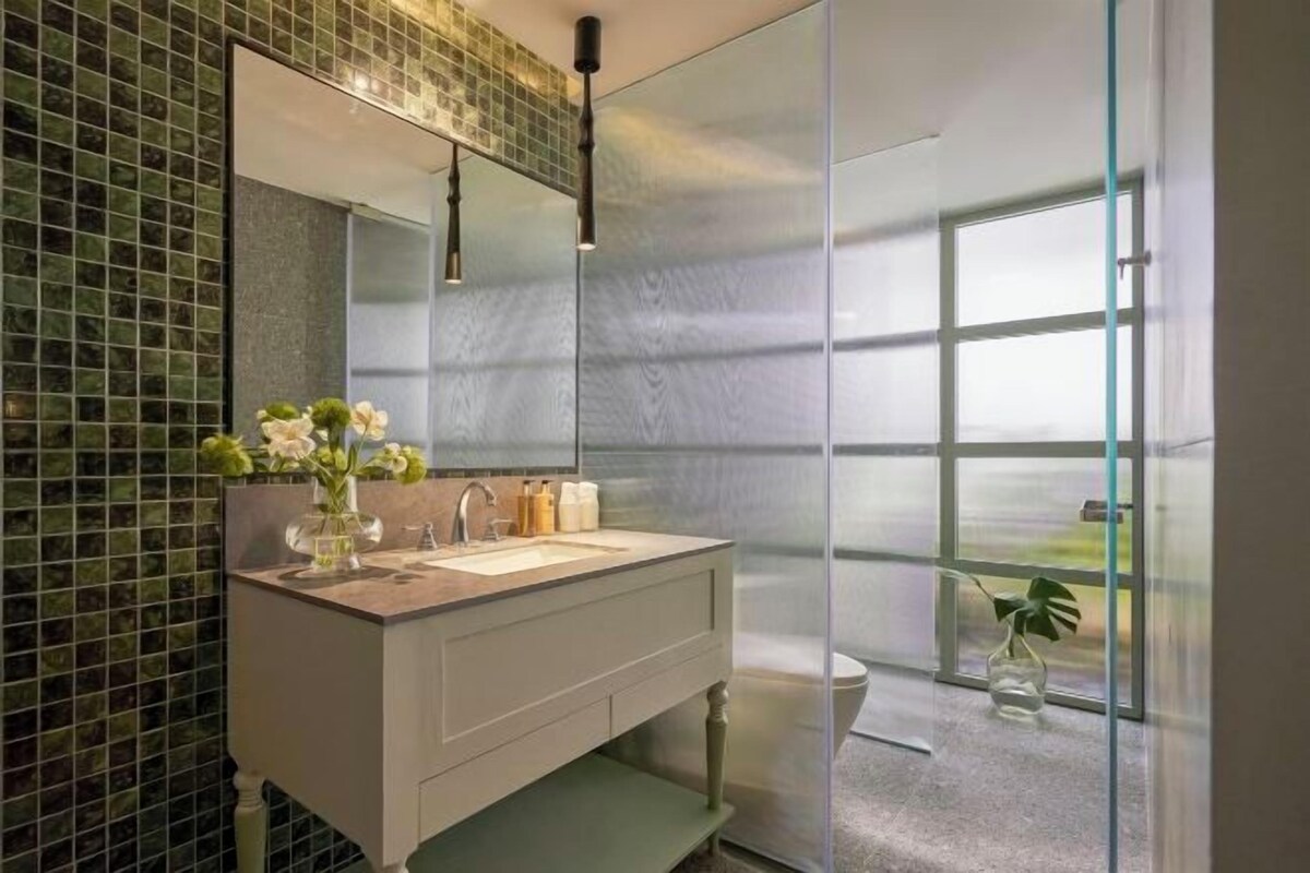 A modern bathroom with a large mirror, a white vanity, and a view of a green landscape through a glass door.