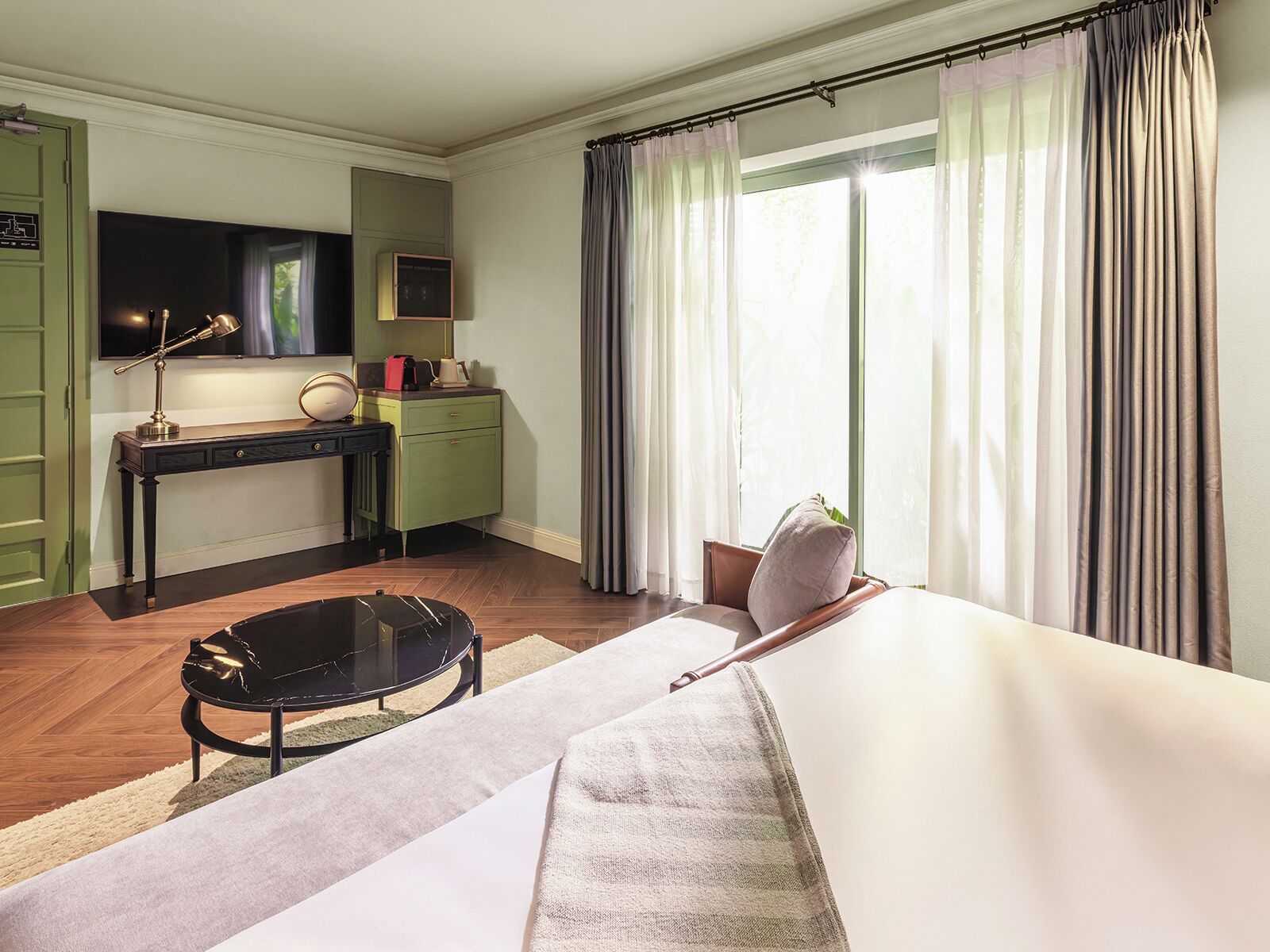 A hotel room with a bed, a desk with a lamp, a chair, a suitcase, and a TV on the wall.