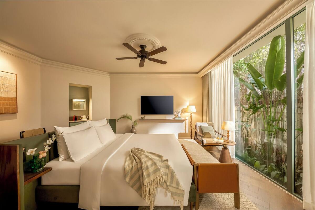 A hotel room with a bed, a TV, a desk, a chair, a ceiling fan, and a large window with a view of greenery.