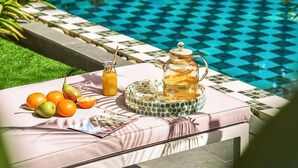 Escena junto a la piscina con una mesa dispuesta para té y fruta, rodeada de exuberante vegetación.