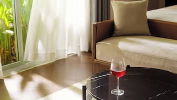A room with a bed, a round table with a glass of red wine, and a view of greenery through a window.