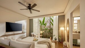 Habitación de hotel con cama, televisión, ventilador de techo, ventana con cortinas y baño visible a través de una puerta abierta.