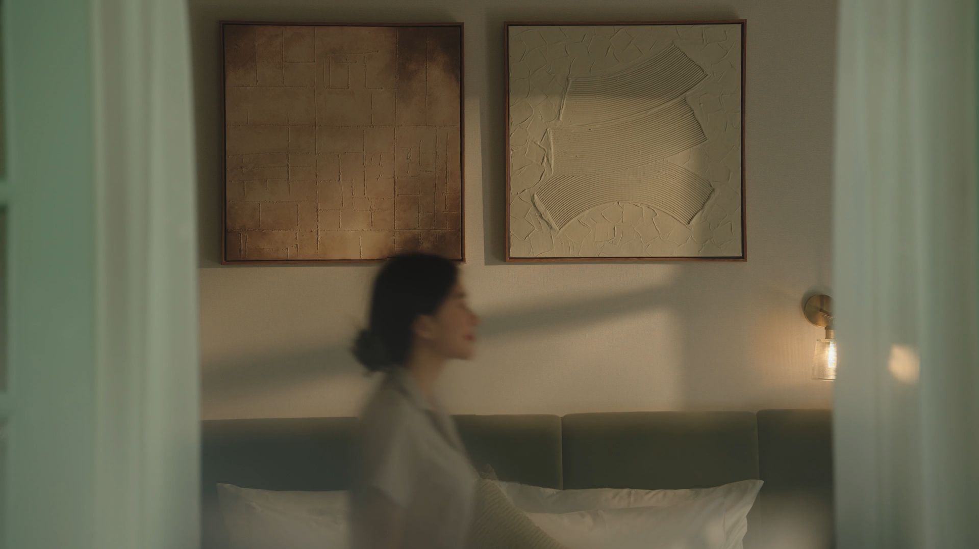 A room with a bed, a wall-mounted light, and two abstract paintings.