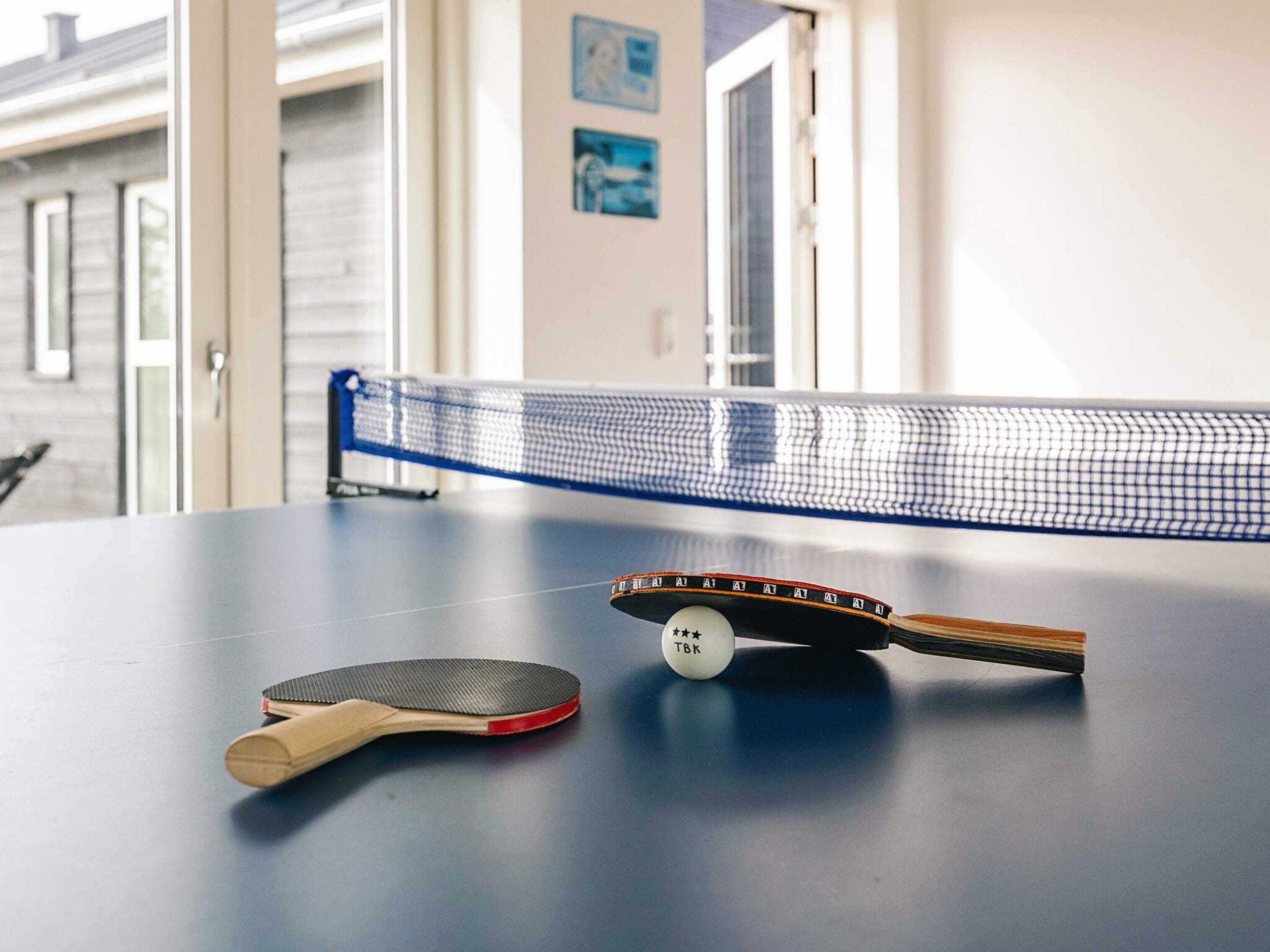 A ping pong table with a net, a paddle, and a ball in a room with a door and a window.