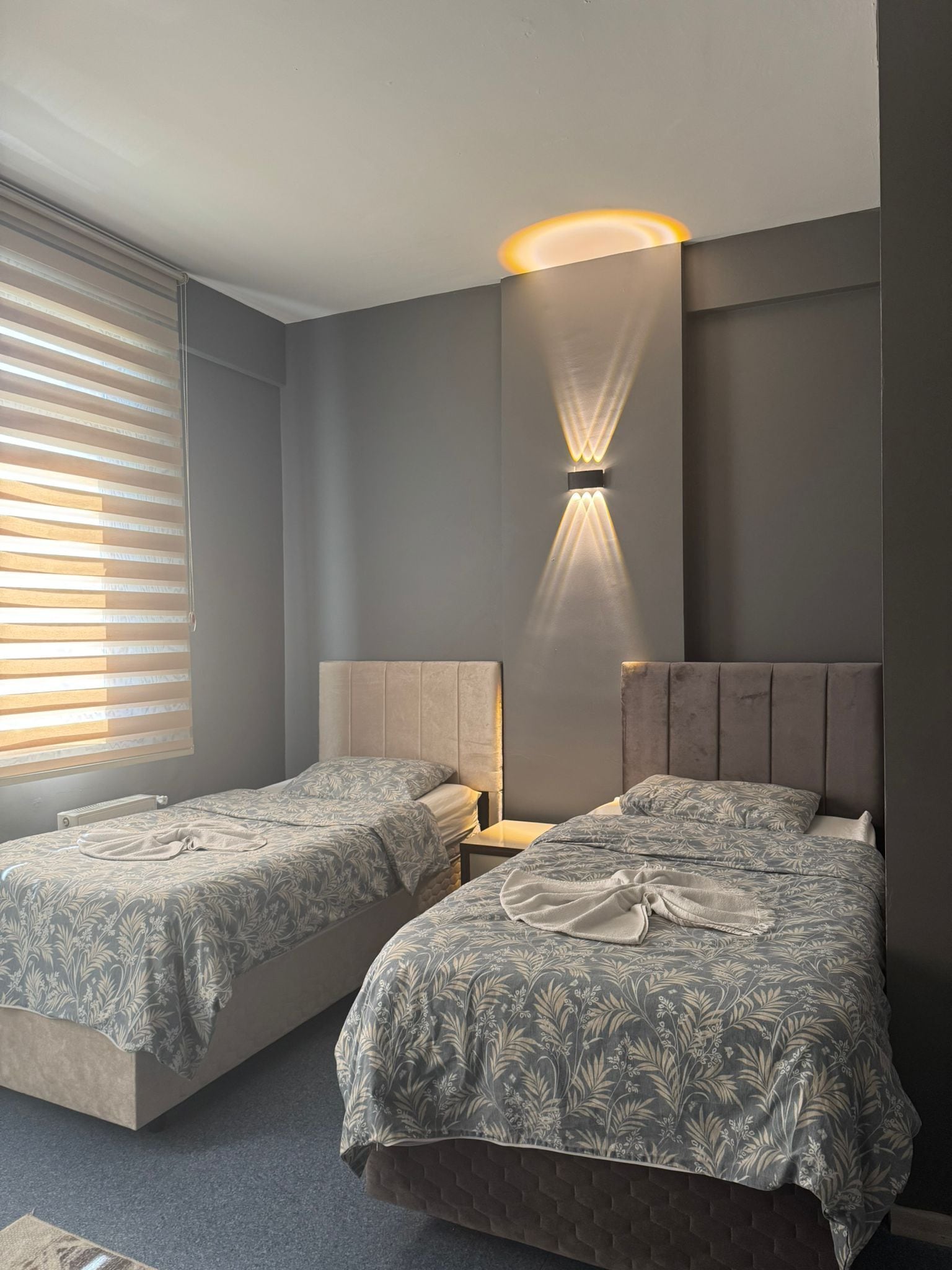 A bedroom with two beds, a wall-mounted light fixture, and a window with blinds.