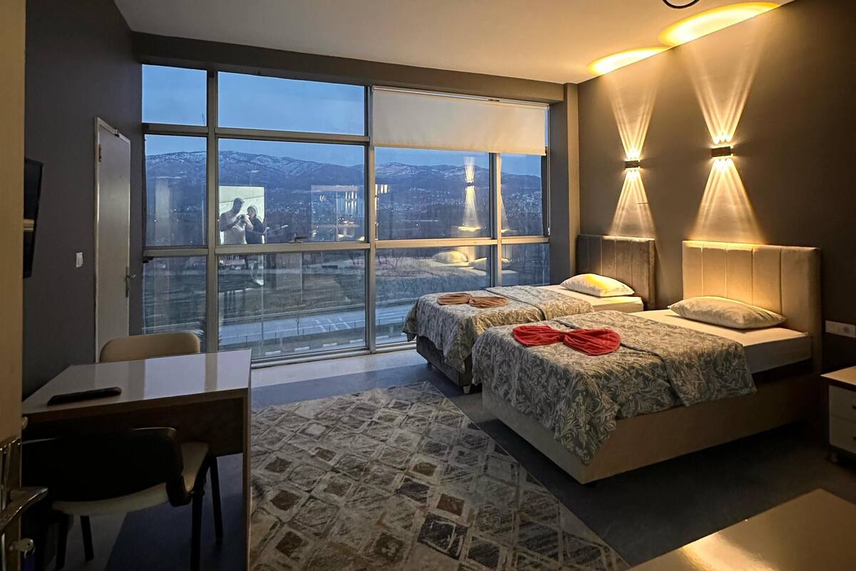 A modern bedroom with a large window, two bedside lamps, a desk, and a patterned rug.