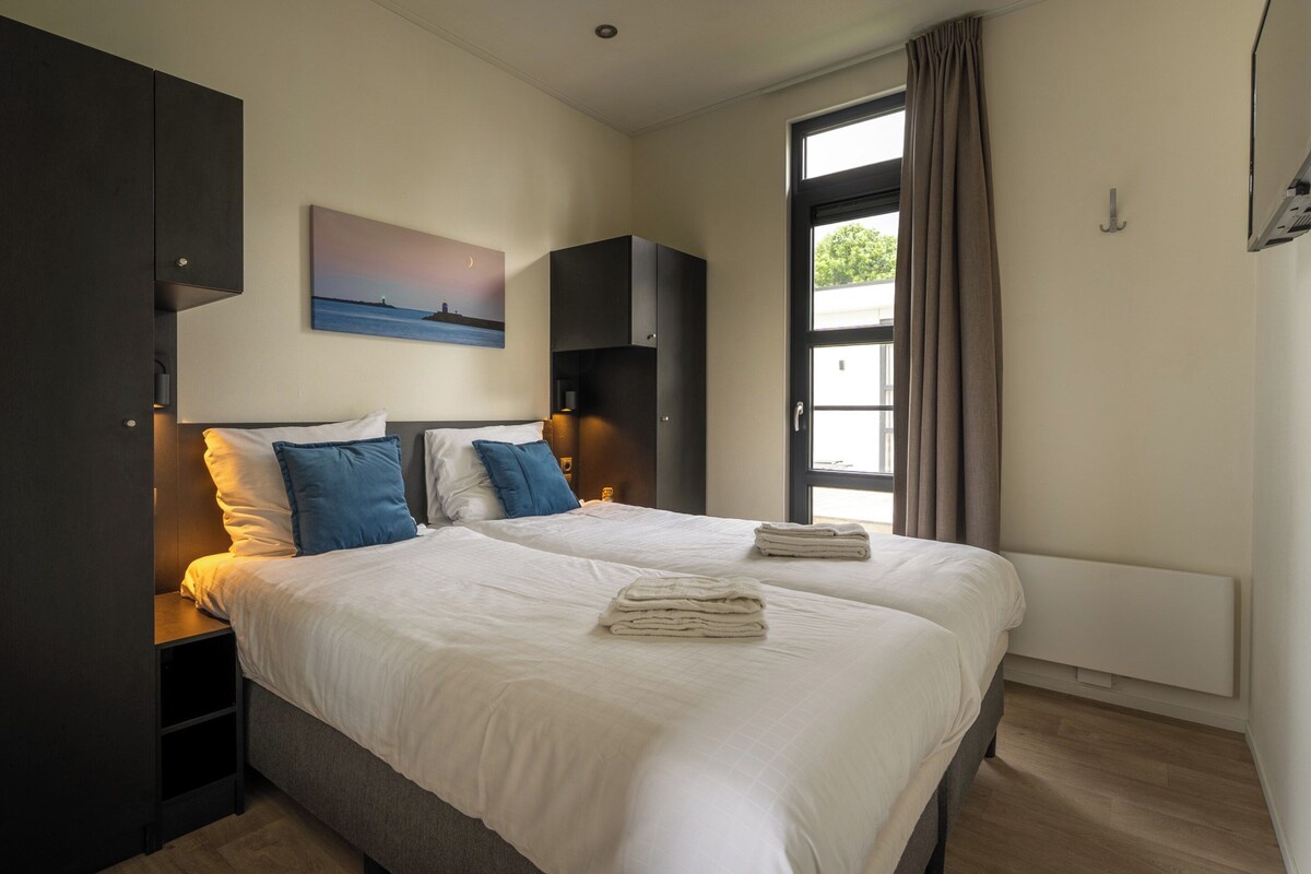 Une chambre d’hôtel avec un grand lit, une table de chevet, une lampe fixée au mur, un tableau et une fenêtre avec des rideaux.