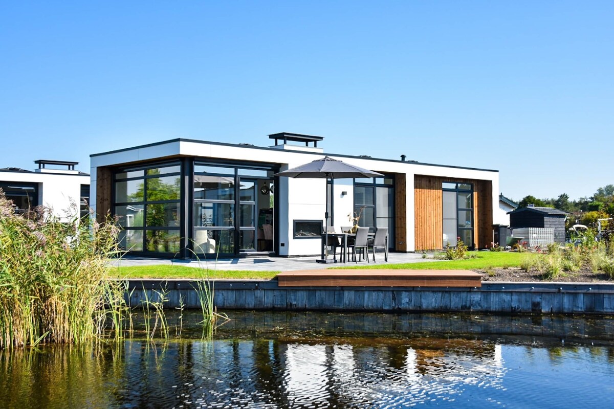 A modern house with large windows, a patio with outdoor furniture, and a pond in the foreground.