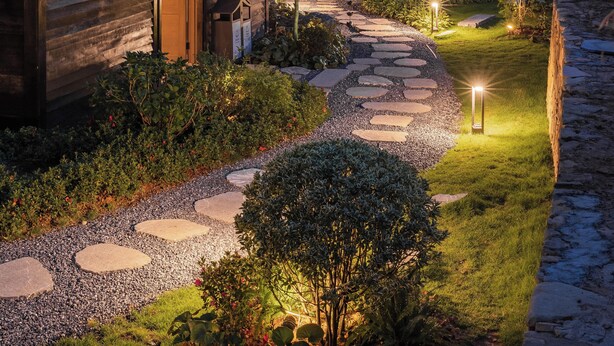 Una casa moderna con un camino de piedra que lleva a la entrada, rodeada de abundante vegetación e iluminada por una luz ambiental.