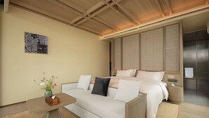 Una habitación de hotel moderna con sofá, una mesita de noche con un jarrón de flores, una cama con almohadas y un ventanal grande.