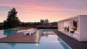 Zona de piscina con terraza de madera, sillas de descanso y mesas dispuestas para comer, con vistas a un jardín ajardinado y colinas al fondo.
