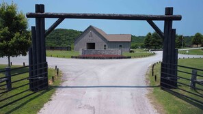 A rural road leading to a wooden barn with a stone foundation, surrounded by grassy fields and trees.
