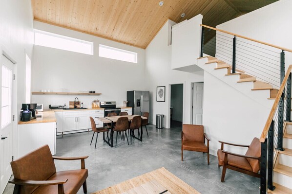 A spacious living area with a dining table, chairs, a kitchen with white cabinets, and a wooden staircase.