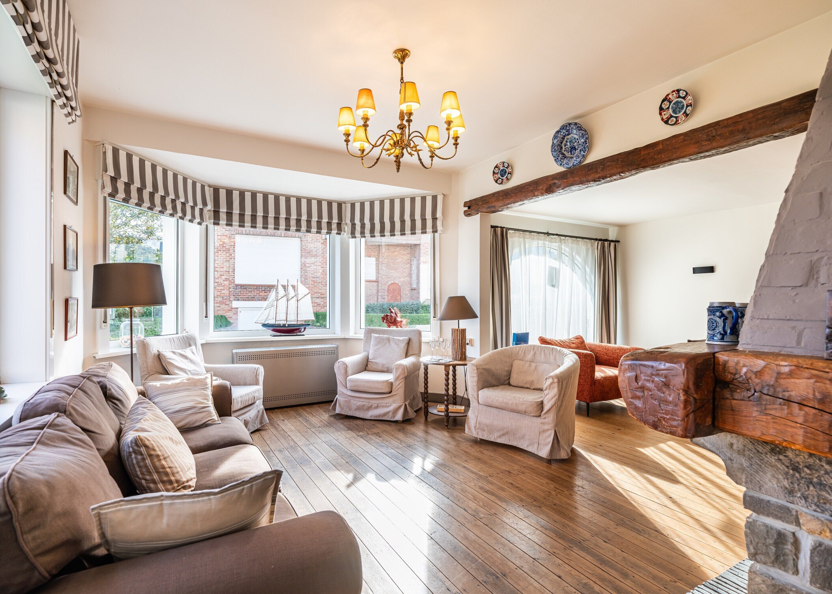 A spacious living room with a wooden floor, a chandelier, and a view of a brick building through large windows.