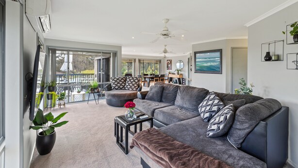 A modern living room with a large gray sectional sofa, a small black coffee table, and a brown ottoman. There is a large window with a view of the outdoors, a ceiling fan, and a painting on the wall. - Sapphire Belle: Views and boat Parking (Eden)