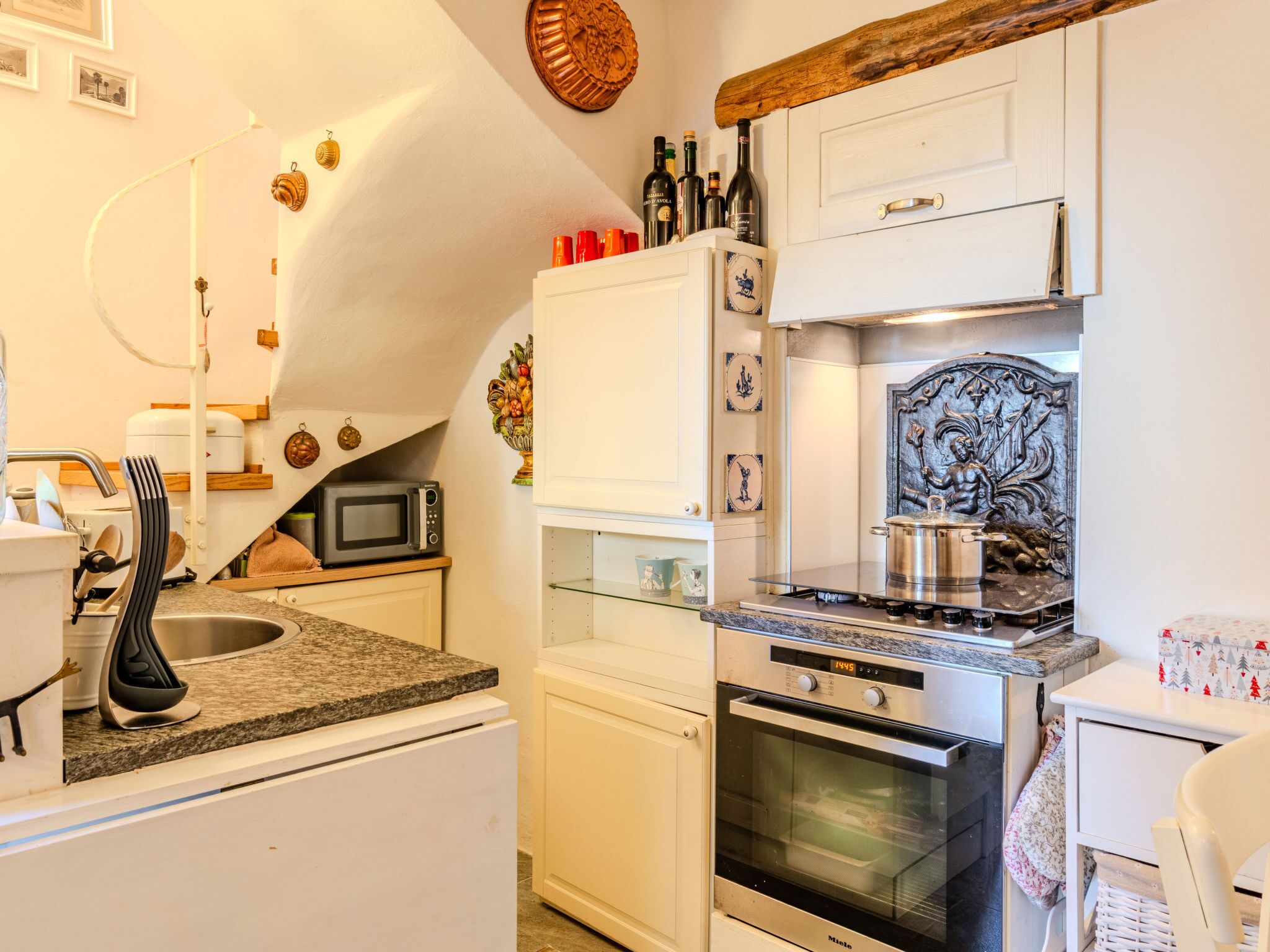 Cozinha compacta com armários brancos, micro-ondas, forno e pia. Há pratos decorativos na parede e uma viga de madeira acima dos armários.