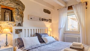 Een slaapkamer met een stenen muur, een wit bed met beddengoed met patroon, twee bedlampen en een raam met witte gordijnen.