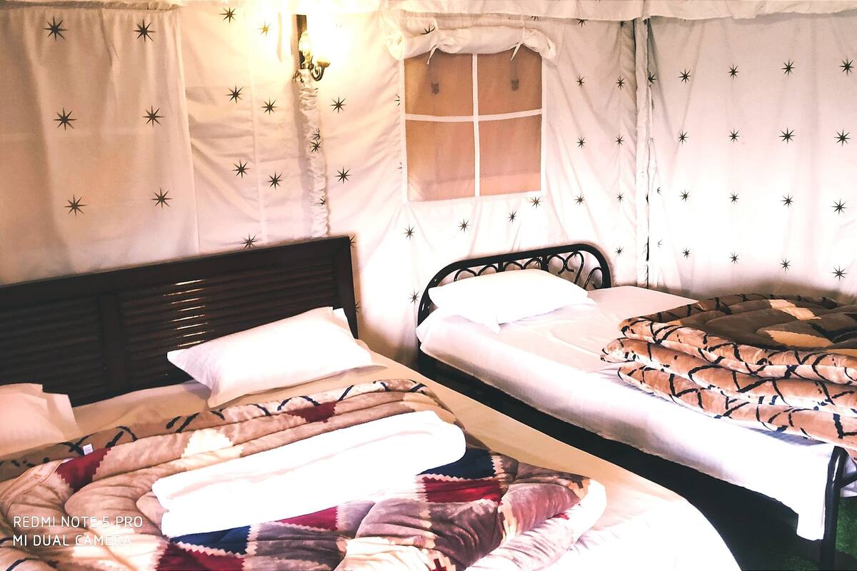 A room with two beds, each with a patterned blanket. There is a window with a curtain, and a wall-mounted light fixture.