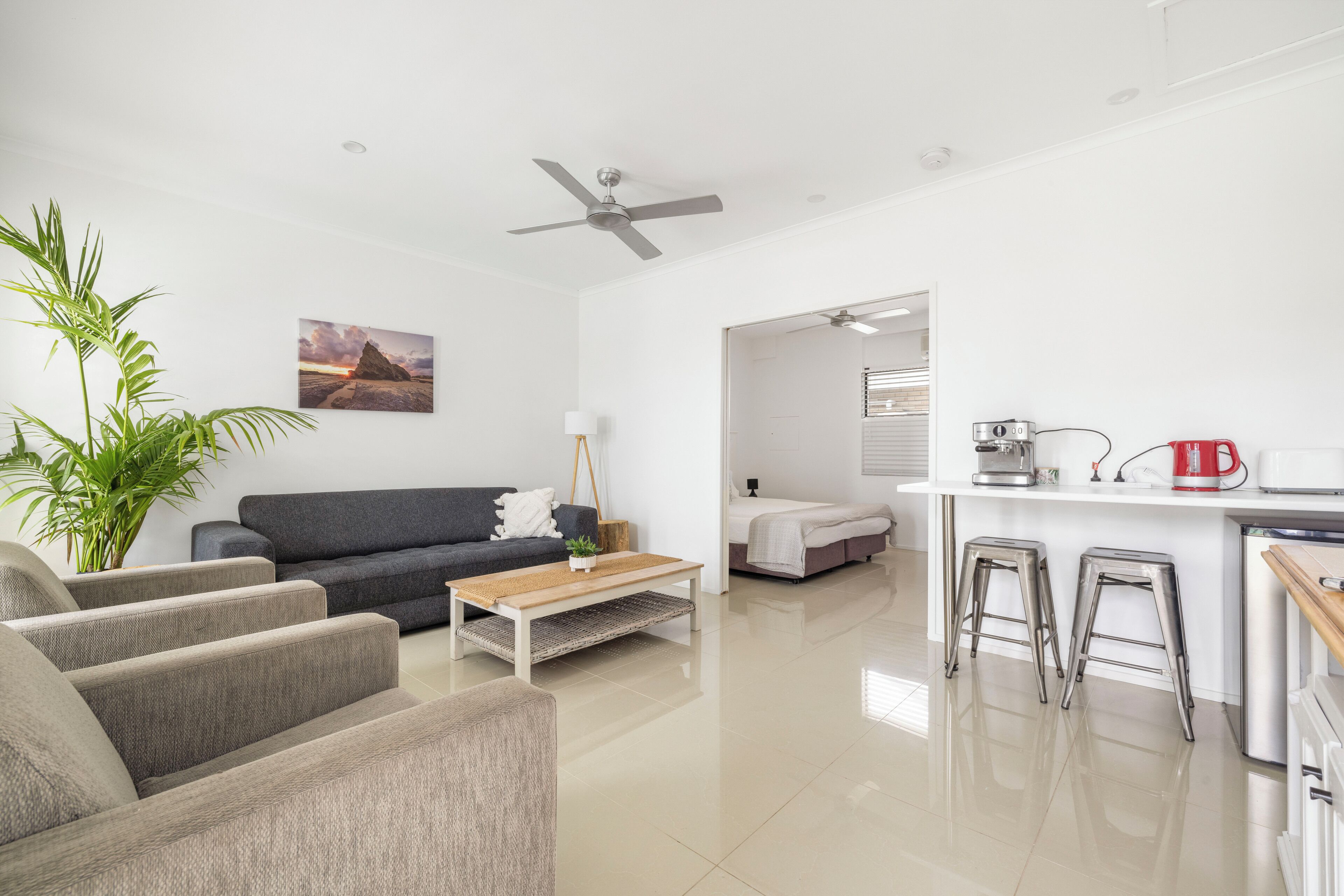 A modern living room with a grey sofa, a wooden coffee table, and a large plant. There is a kitchen area with a white countertop and bar stools.