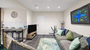 A modern living room with a grey sofa, a coffee table, and a TV. There is a kitchen area with a white tiled backsplash and a dining table with bar stools. A large abstract painting is on the wall, and a door is visible in the background. - 5 Bedroom Oxford Cooper House, Sleep 11, Parking (Oxford)