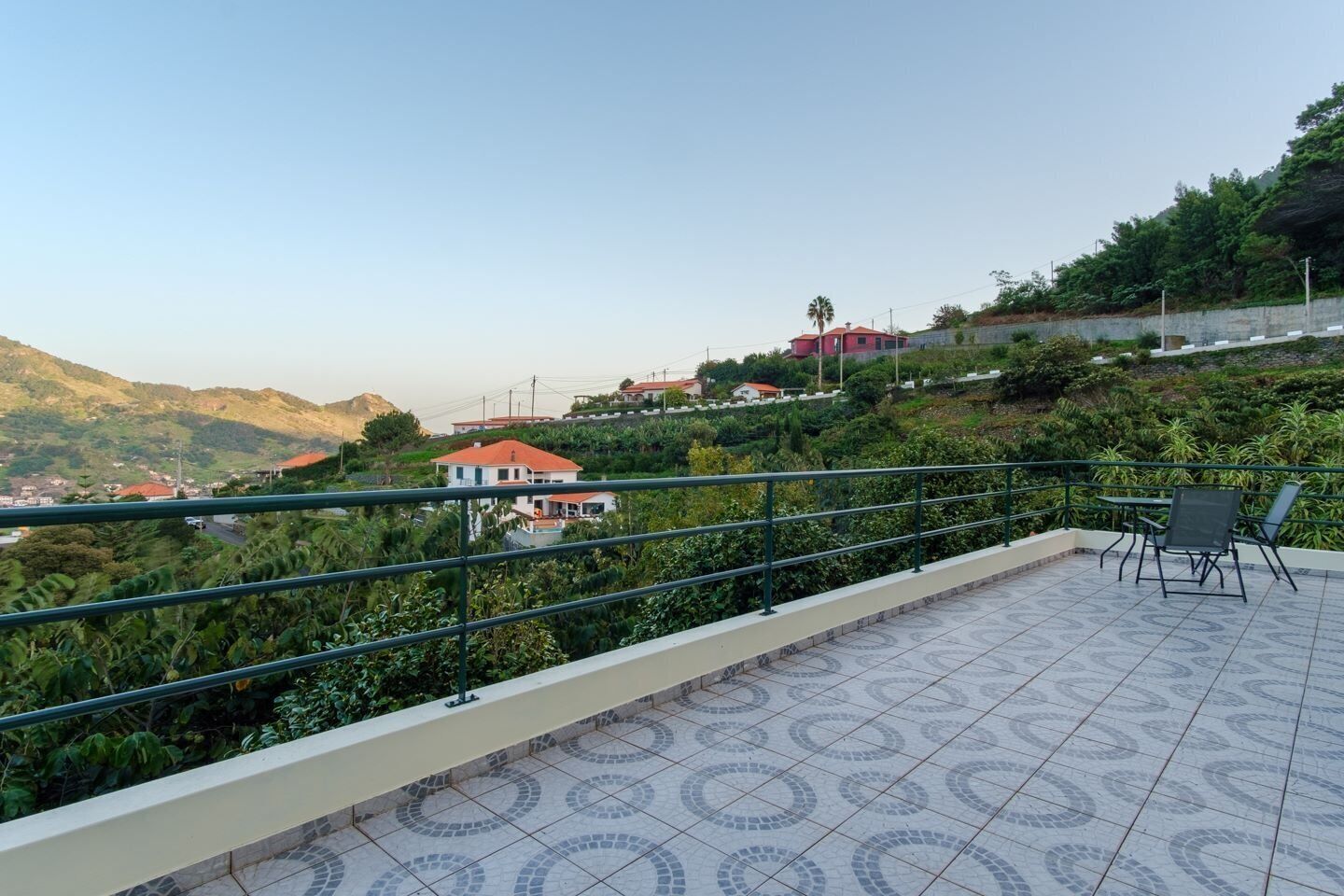 Eine Terrasse mit Fliesenboden, einem Metallgeländer und Blick auf einen Hang mit Gebäuden.